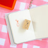 A small wooden rubber stamp with a flower design is shown on an open notebook, having been used to stamp an orange flower mark. The notebook is placed on a pink and white checkered surface, with a red ink pad visible in the top-left corner.