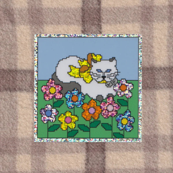 A square patch featuring a pixel-art style illustration of a white cat among colourful flowers on a green field under a light blue sky, bordered by a floral pattern. The patch is placed on a beige and brown plaid fabric background.