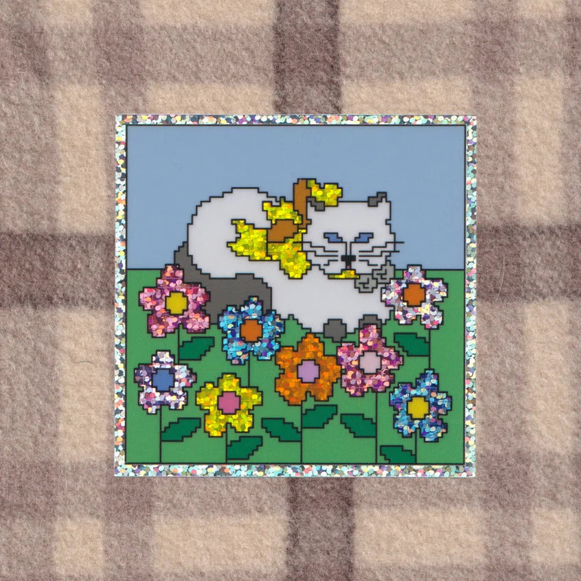 A square patch featuring a pixel-art style illustration of a white cat among colourful flowers on a green field under a light blue sky, bordered by a floral pattern. The patch is placed on a beige and brown plaid fabric background.