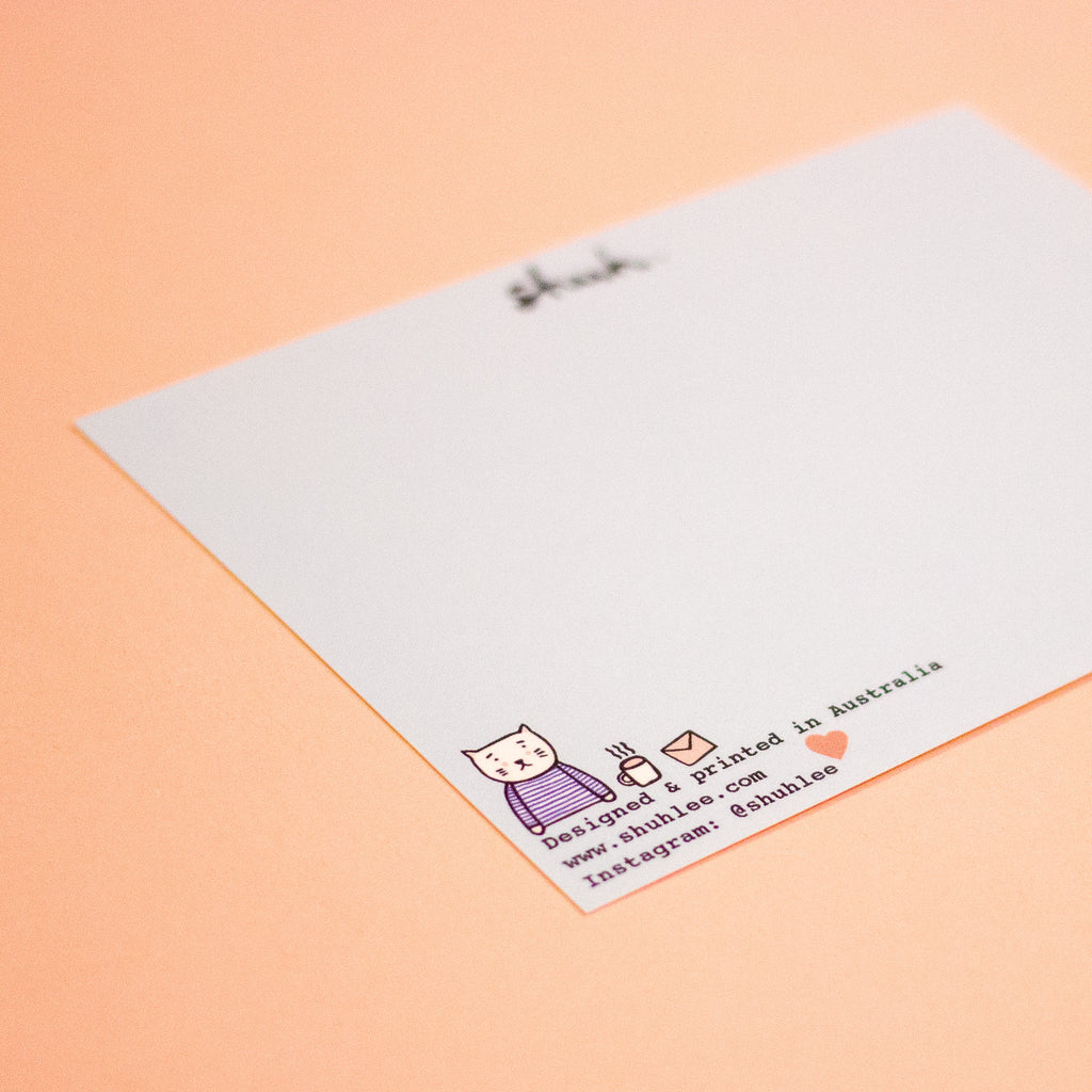 A close-up of a rectangular postcard with a light blue background and maker details on the corner photographed on a peach backdrop.