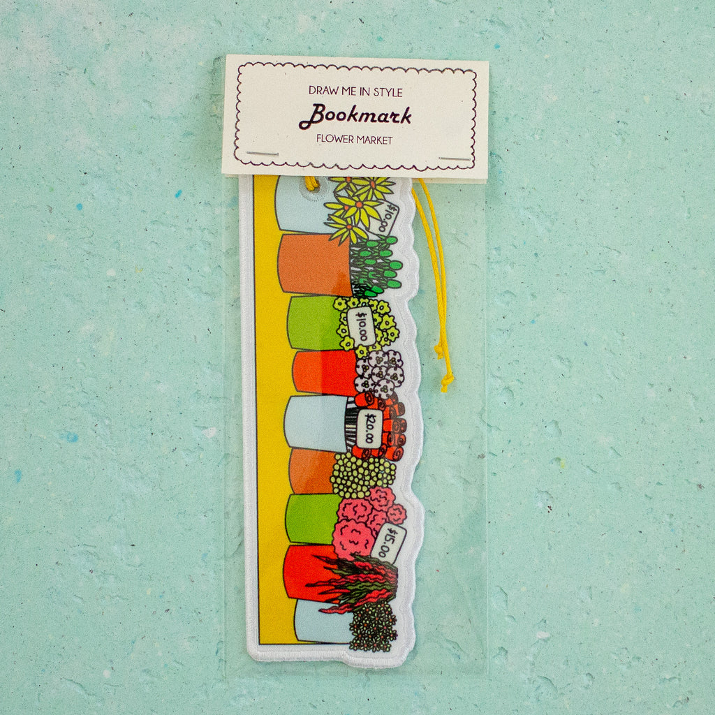 A decorative bookmark shaped like a flower market scene, packaged in a clear sleeve with a white header card that reads 'Draw Me in Style Bookmark - Flower Market.' The bookmark features illustrated flower pots, plants, and a yellow drawstring, shown against a light aqua textured background.