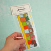 A hand holds a packaged bookmark shaped like a flower market row of colourful pots with illustrated plants and labels, against a pale mint background.
