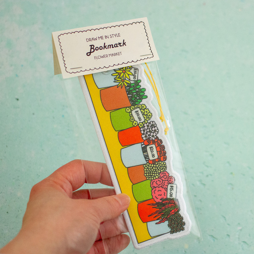 A hand holds a packaged bookmark shaped like a flower market row of colourful pots with illustrated plants and labels, against a pale mint background.