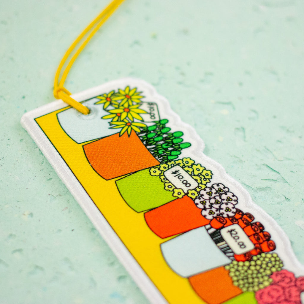 A close-up of a colorful rectangular hanging tag or bookmark with a yellow loop and string, featuring illustrated flower pots and plants on a pale mint background.