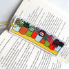 A rectangular acrylic bookmark with a colourful illustrated row of potted plants and small labels, hanging from a yellow string over an open book with printed text.
