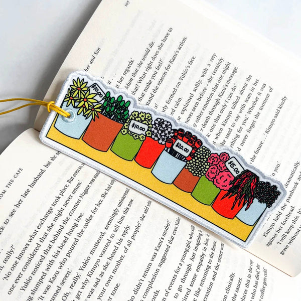 A rectangular acrylic bookmark with a colourful illustrated row of potted plants and small labels, hanging from a yellow string over an open book with printed text.