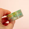 A hand holding a roll of washi tape with a colourful flower pattern against a peach background.