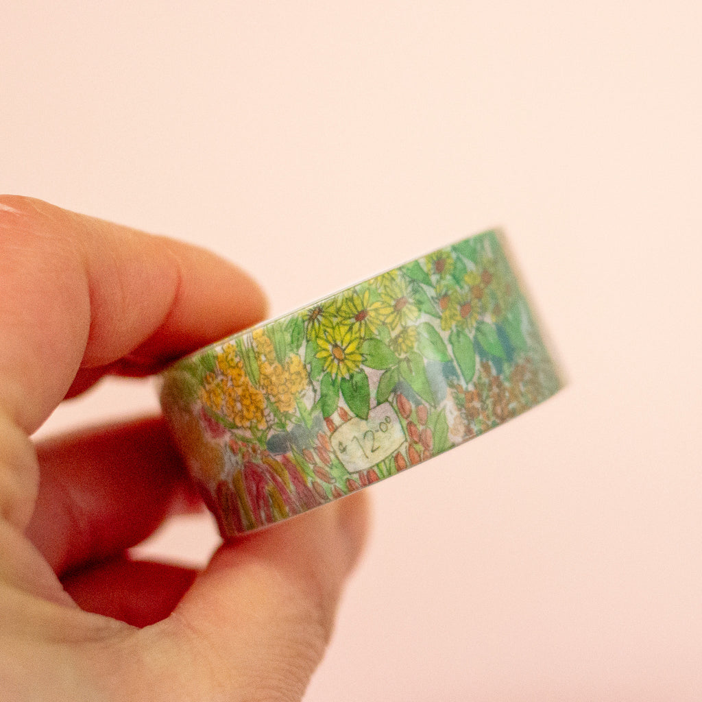 A hand holding a roll of washi tape with a colourful flower pattern against a peach background.