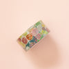 A roll of washi tape with a colourful floral pattern, displayed diagonally on a soft peach-pink background.