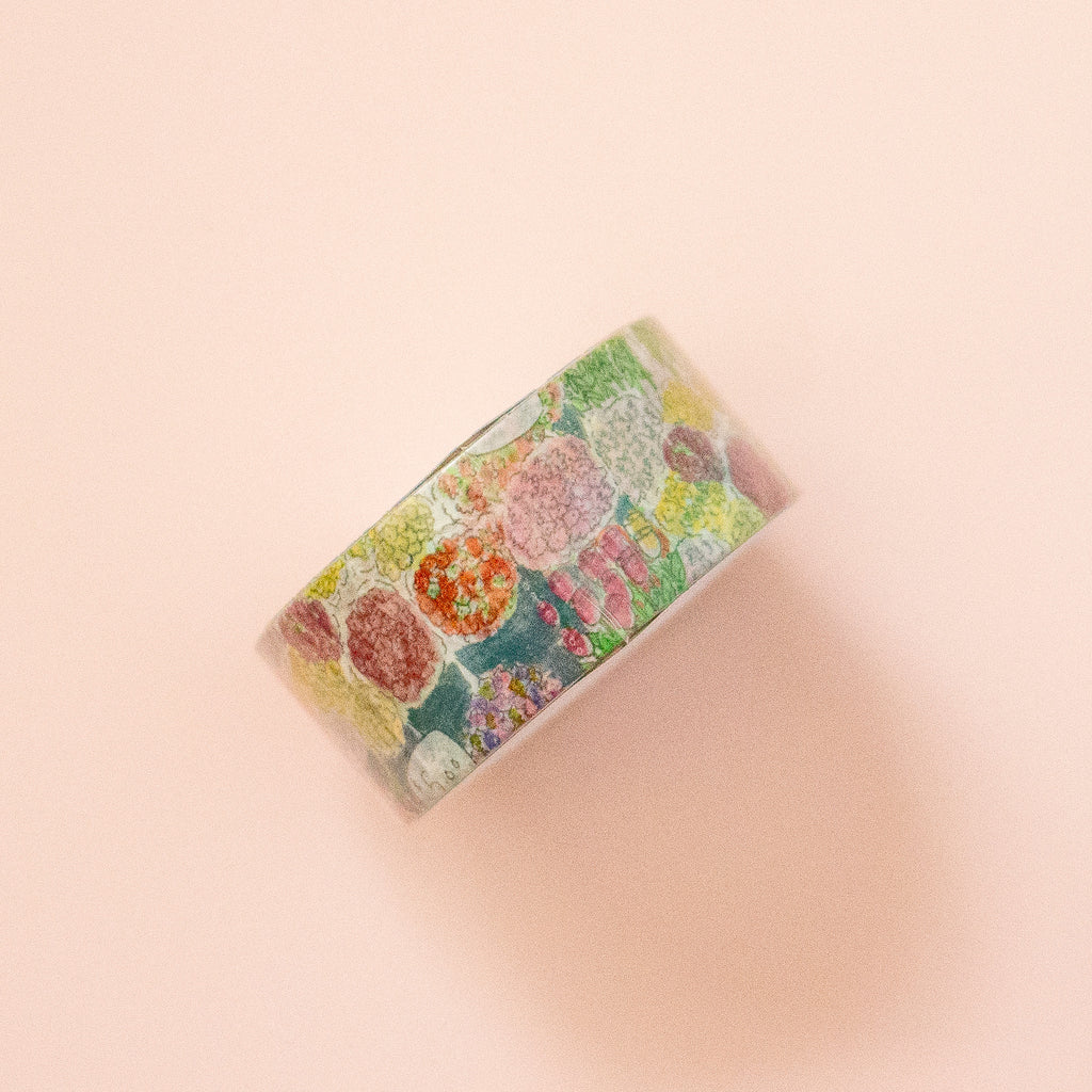 A roll of washi tape with a colourful floral pattern, displayed diagonally on a soft peach-pink background.