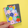 A hand holding a sticker with a colourful floral pattern over a dark grey card, set against a solid yellow background.
