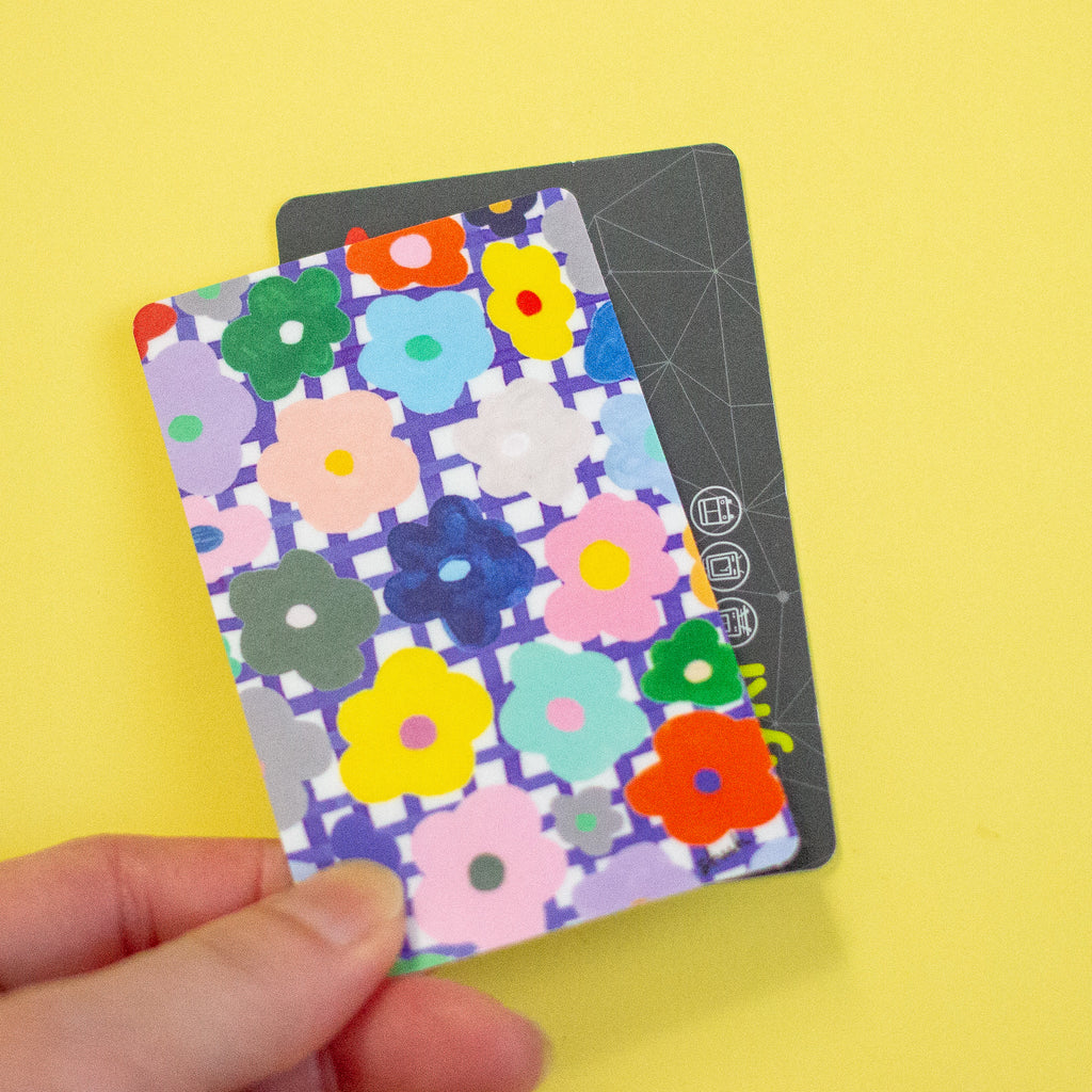 A hand holding a sticker with a colourful floral pattern over a dark grey card, set against a solid yellow background.