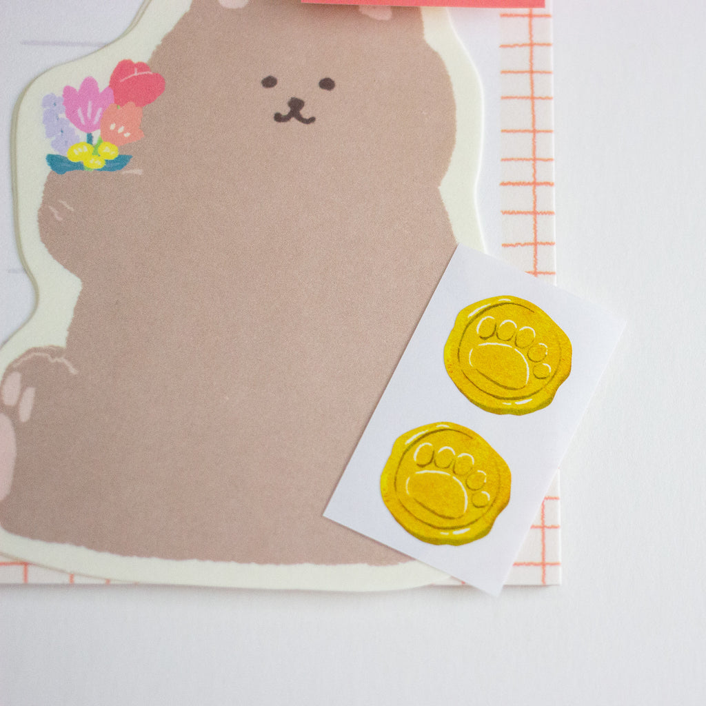 A cute bear-shaped paper with a blank center, decorated with a small flower in the top-left corner, shown alongside a sheet of two yellow paw-print wax seals on a white background.