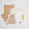 A stationery set featuring a cute teddy bear sticker, two brown kraft envelope, and grid writing paper with pink grid borders and gold seal stamp sticker, arranged on a light grey background.