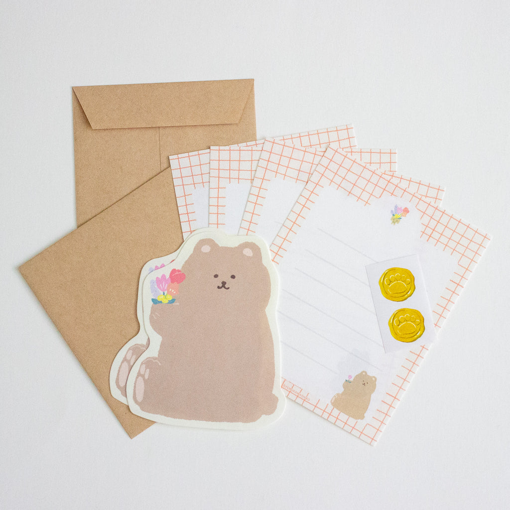 A stationery set featuring a cute teddy bear sticker, two brown kraft envelope, and grid writing paper with pink grid borders and gold seal stamp sticker, arranged on a light grey background.