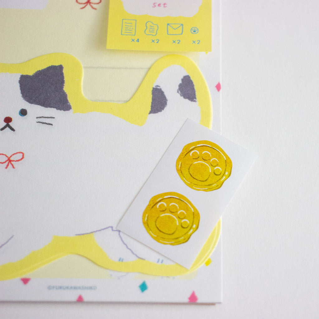 A close-up of a stationery set featuring a white card with a yellow cat outline and a small white sticker sheet with two yellow paw-print designs, set against a white background.