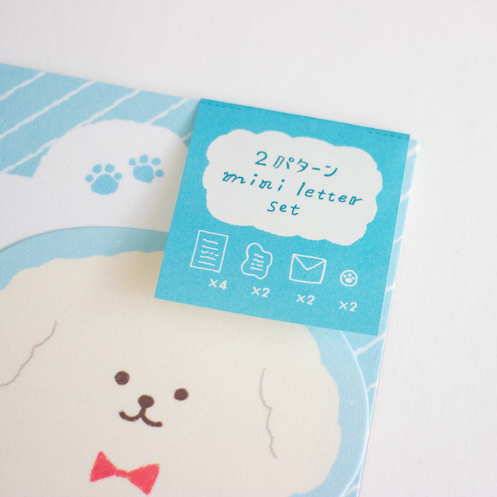 A close-up of a cute stationery set featuring a printed package card for a 'mini letter set' in light blue, placed over a matching printed sheet with a white dog face and red bow tie.