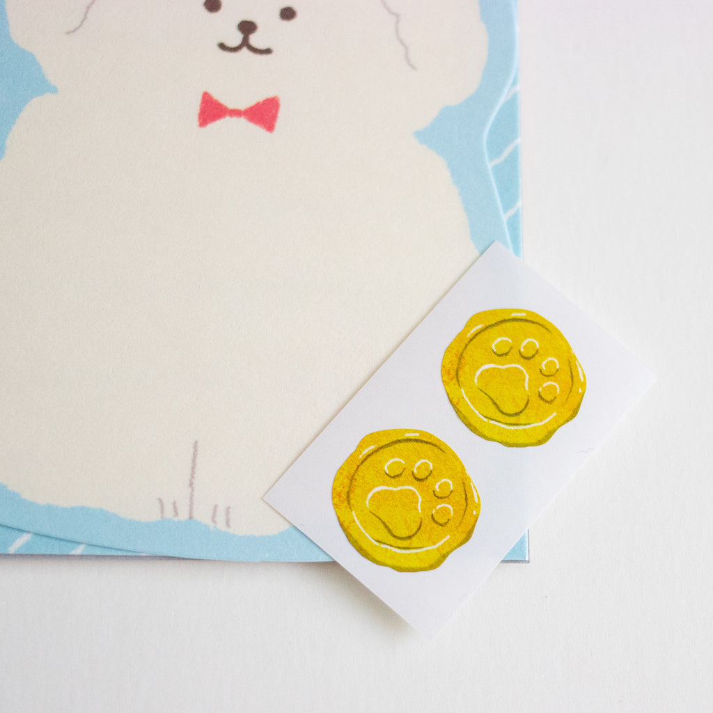 Two round yellow paw-print stickers on a small white card, placed on top of a white surface with a partially visible illustrated card featuring a white dog and red bow.