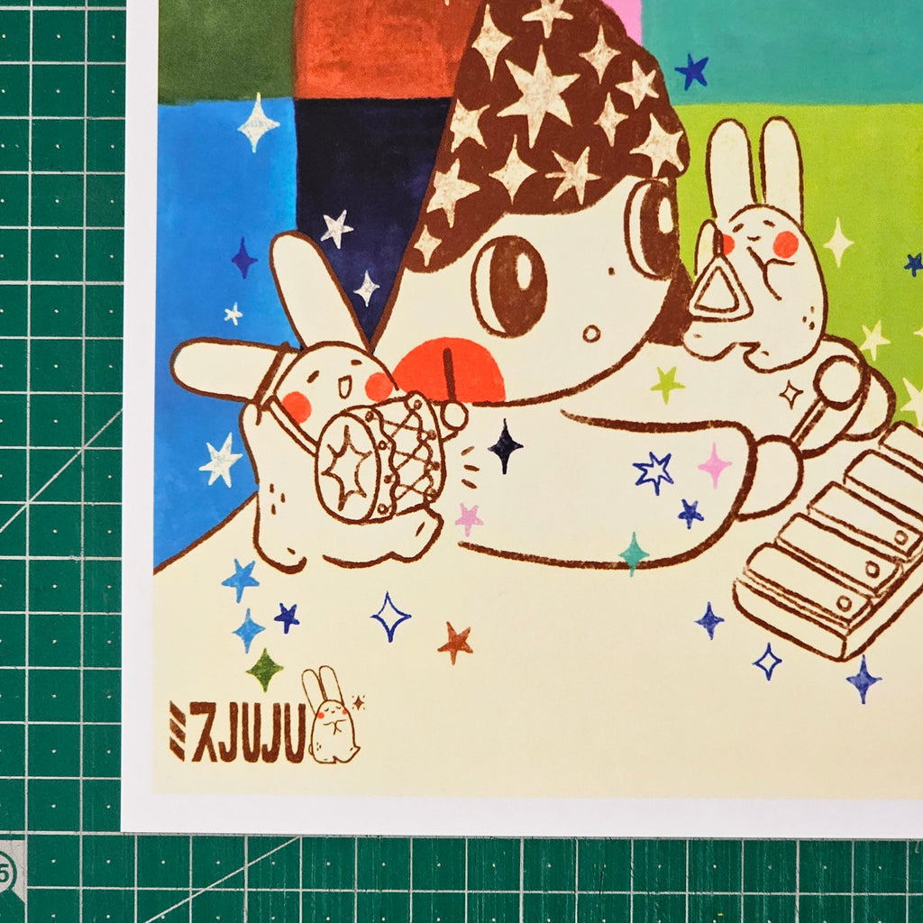 An art print featuring a whimsical cartoon illustration of a character with star-patterned hair holding a star-laden cup, with two rabbits and a keyboard on a colourful background. The print is photographed on a green cutting mat.