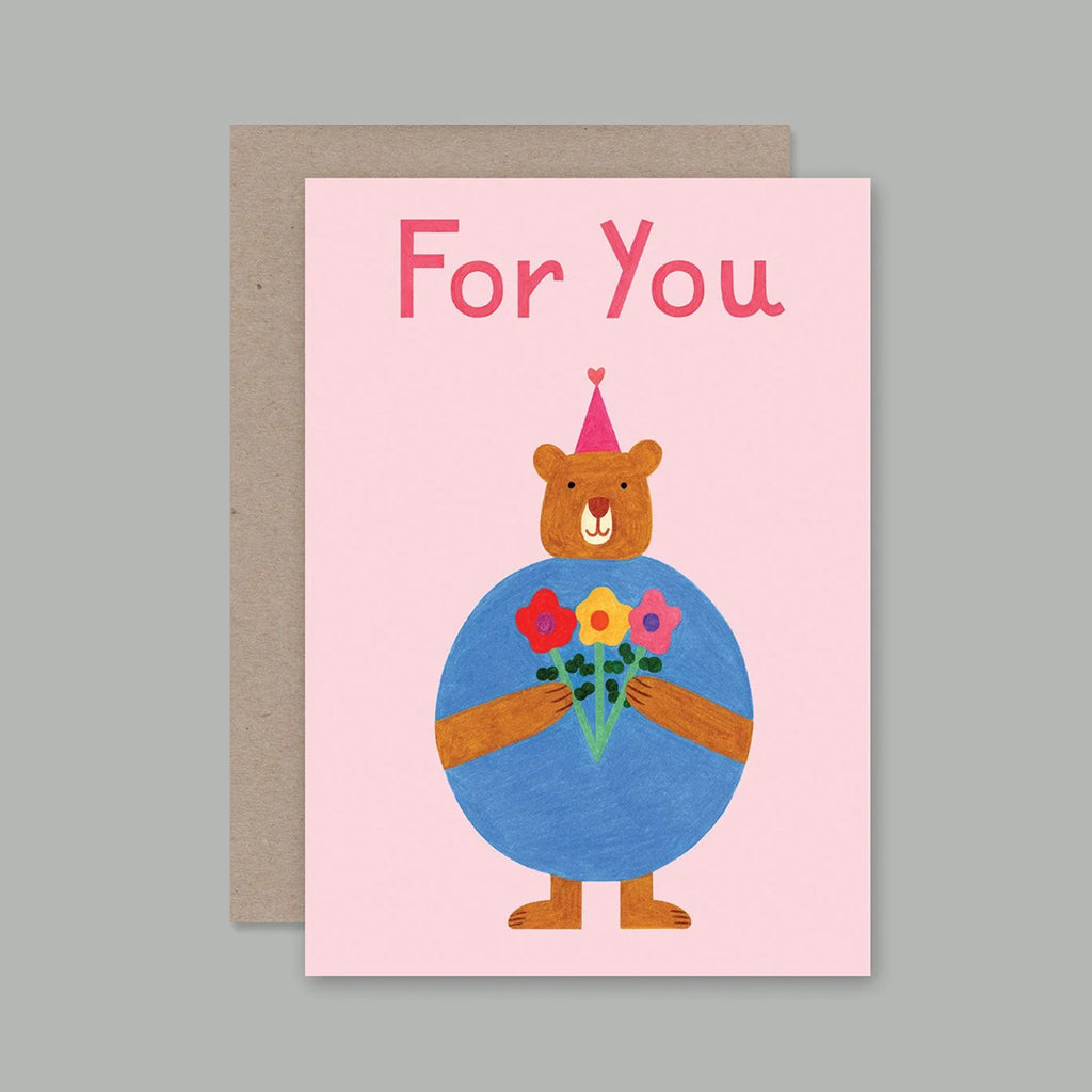 A pink greeting card with the words 'For You' in red lettering, featuring a cartoon bear wearing a party hat and holding a bouquet of colourful flowers. The card is shown with a brown envelope behind it on a flat grey background.