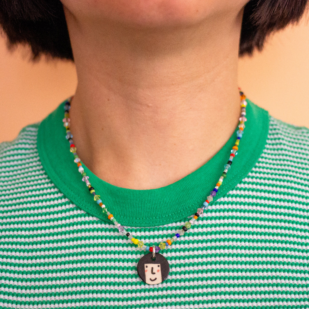 Person wearing a colourful beaded necklace with a small round pendant on a green and white striped shirt.