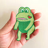 Green frog patch with a red heart held in a hand against a light background