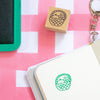 A wooden rubber stamp with a design of a basket containing fruits, shown next to a green ink pad and a notebook with a green stamped impression on pink and white checkered paper.