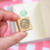 A hand holding a small wooden stamp block featuring a black ink illustration of a basket with a fruits inside, against a soft pink and white background.
