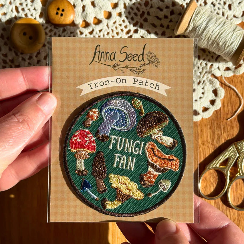Iron-on patch with 'Fungi Fan' design held by a hand, on a wooden surface with sewing tools.