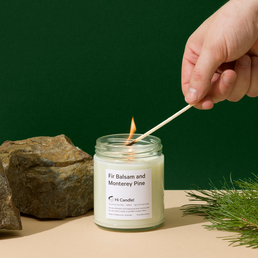 Hand lighting a candle with a match, surrounded by stones and greenery on a beige surface.