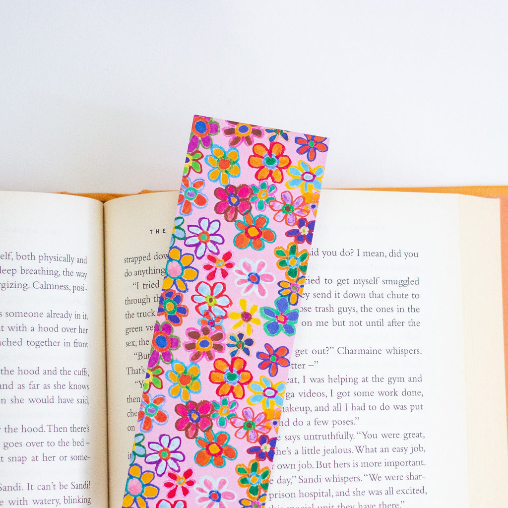 A colourful floral-patterned bookmark with pink, orange, yellow, green, and blue flowers, shown resting on an open book against a plain white background.