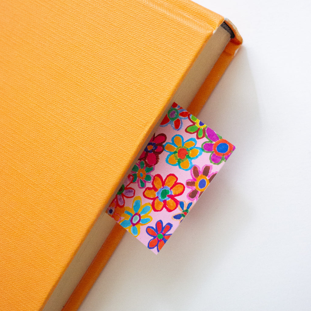 A close-up of an orange hardcover notebook or book with a small rectangular paper bookmark or page marker tucked between the pages. The bookmark has a pink background with a colourful floral pattern in orange, yellow, green, blue, and red, set against a clean white background.