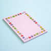 A pink grid notepad with a colourful floral border, photographed on a light blue background.
