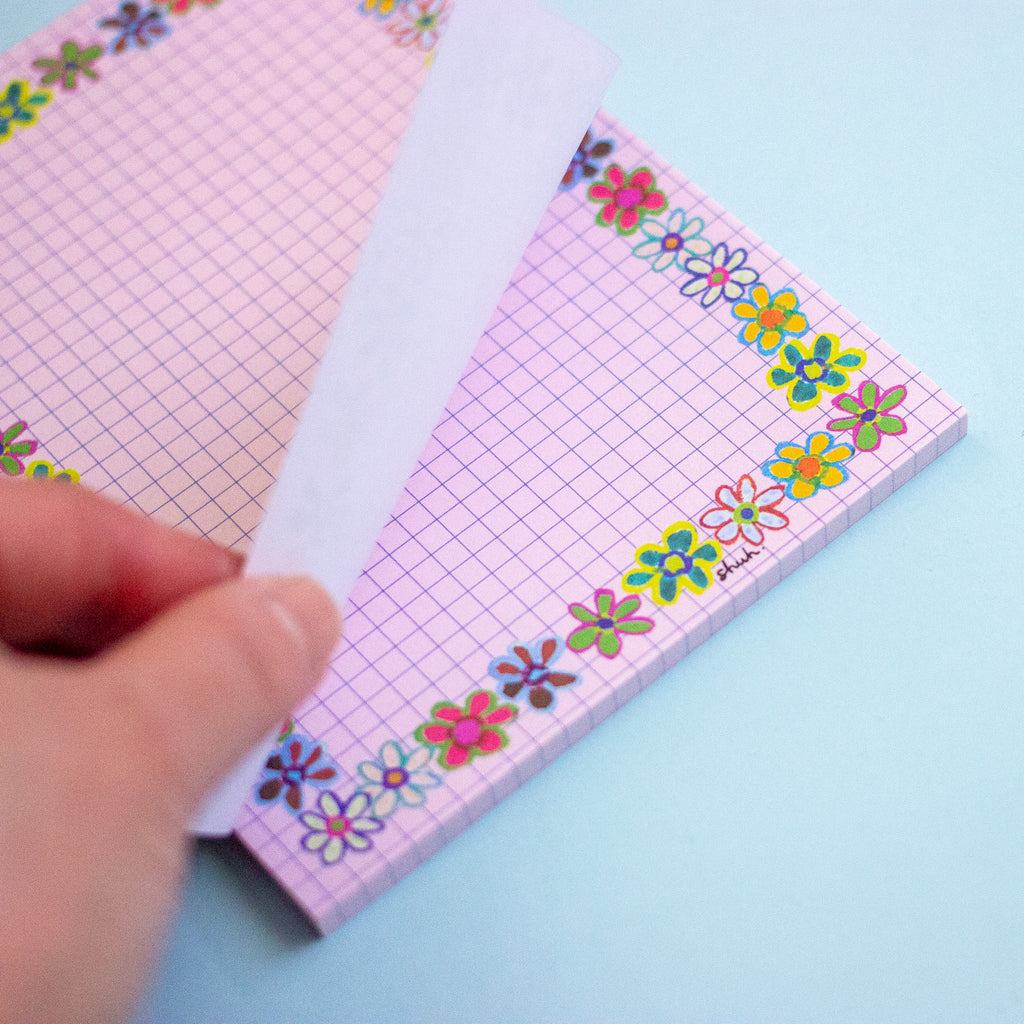 A close-up of a hand peeling back the front page of a pink grid notepad decorated with colourful floral borders, set against a light blue background.
