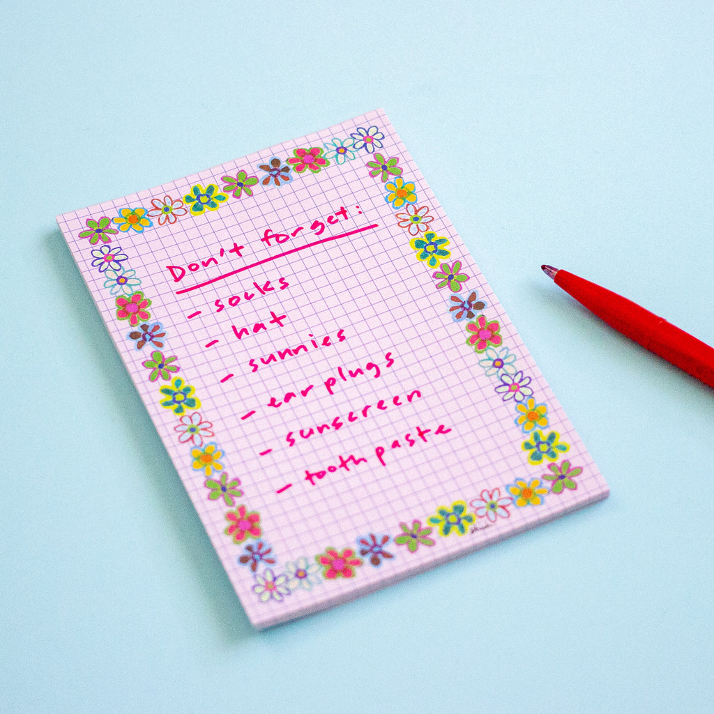 A pink grid notepad with a colourful floral border and handwritten checklist reading 'Don't forget: socks, hat, sunnies, ear plugs, sunscreen, tooth paste' lies on a light blue background next to a red pen.