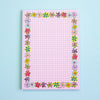 A pink graph paper notepad with a colourful border of small flowers, photographed flat against a light blue background.