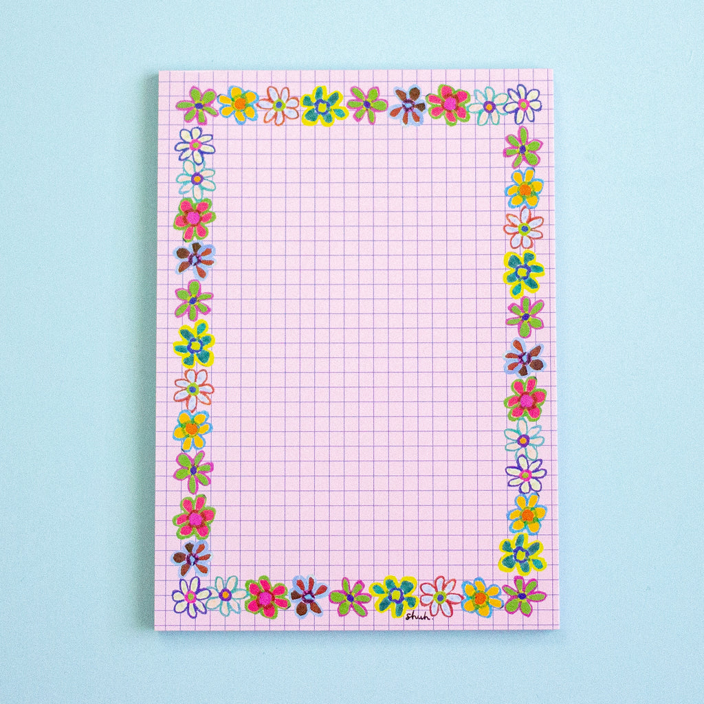 A pink graph paper notepad with a colourful border of small flowers, photographed flat against a light blue background.