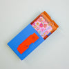 Floral keychain in an open blue box with an orange flap on a light grey background