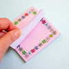 A hand holds a pink notepad with a colourful floral border pattern, showing the white page backing, against a light blue background.