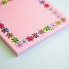 A pink notepad with a colourful floral border, photographed on a light mint-green surface.