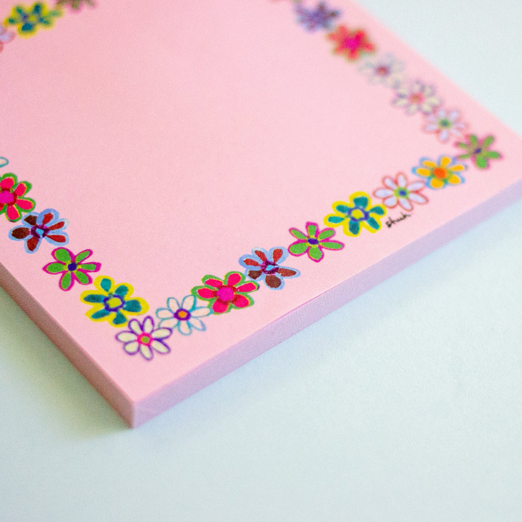A pink notepad with a colourful floral border, photographed on a light mint-green surface.