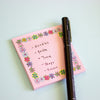 A pink sticky note pad with a colourful floral border and handwritten list items, placed on a pale blue background with a black pen resting on top.