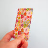 A hand holding a slim rectangular card with a colourful floral pattern on a light pink background, photographed against a plain light blue wall.