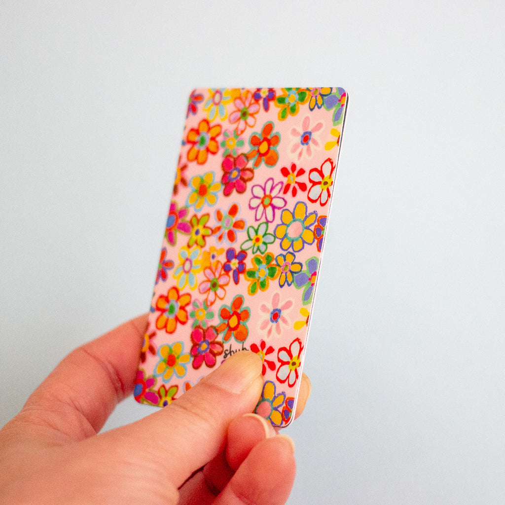 A hand holding a slim rectangular card with a colourful floral pattern on a light pink background, photographed against a plain light blue wall.