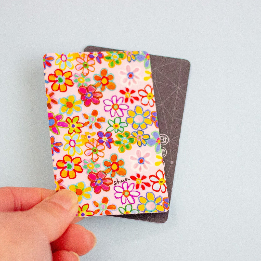 A hand holding a sticker with a colourful floral print over a dark grey card, against a light blue background.