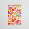 A rectangular sticker with a vibrant, multicoloured floral pattern on a light pink background, photographed flat against a neutral light blue background.