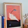 A framed art print featuring a whimsical white bird with a long blue neck and red beak, set against a pink sky and dark ground, styled on a wooden shelf next to books and a small vase.