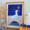 A framed art print featuring a stylised white goose on a dark blue background, styled on a wooden tabletop next to a vase of dried flowers and a white penguin toy.