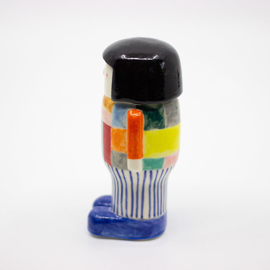 Side view of a handmade ceramic figurine with a black bob hairstyle, colourful patchwork shirt, striped pants, and blue shoes. Designed by shuh.