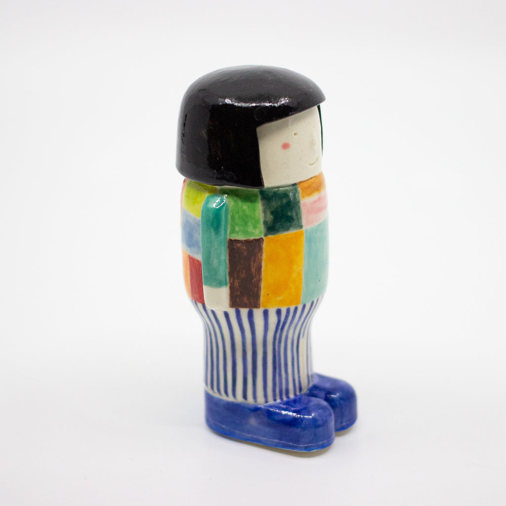 Side view of a handmade ceramic figurine with black bobbed hair, a colourful patchwork top, vertical blue-striped pants, and blue shoes. Designed by shuh.